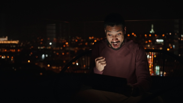 A young African AMerican man sitting on balcony with urban view and using tablet at night