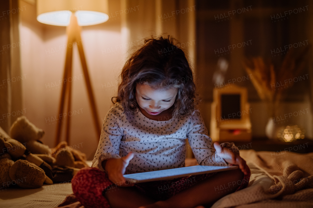 A happy little multiracial girl using tablet on bed in evening at home.