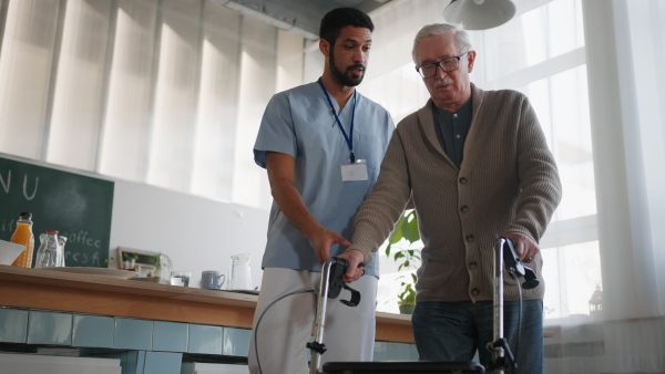 A caregiver helping senior man to walk with walker indoors in retirement nursing hme.