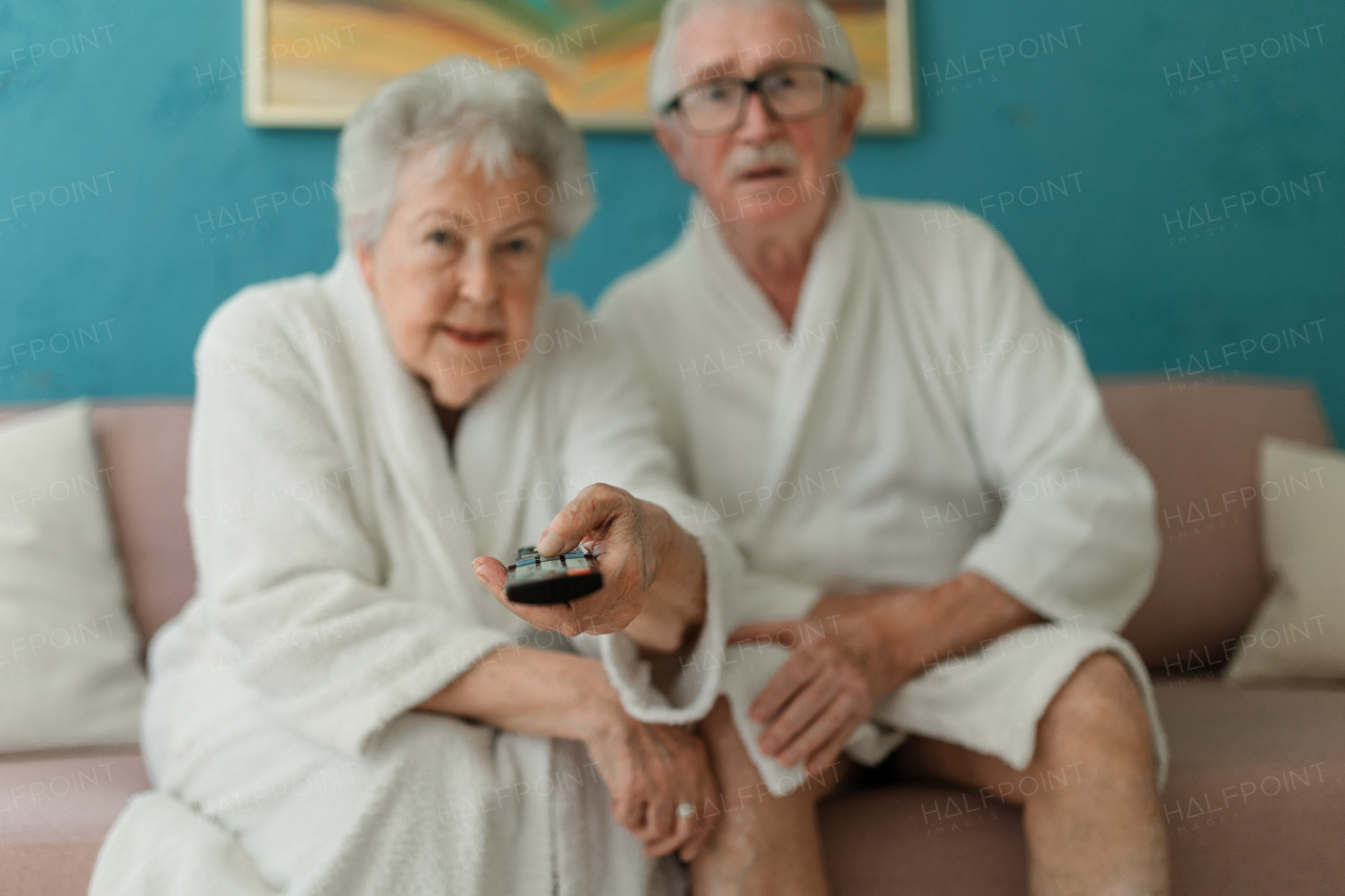 Happy senior couple sitting at a sofa in bathrobes and watching TV.