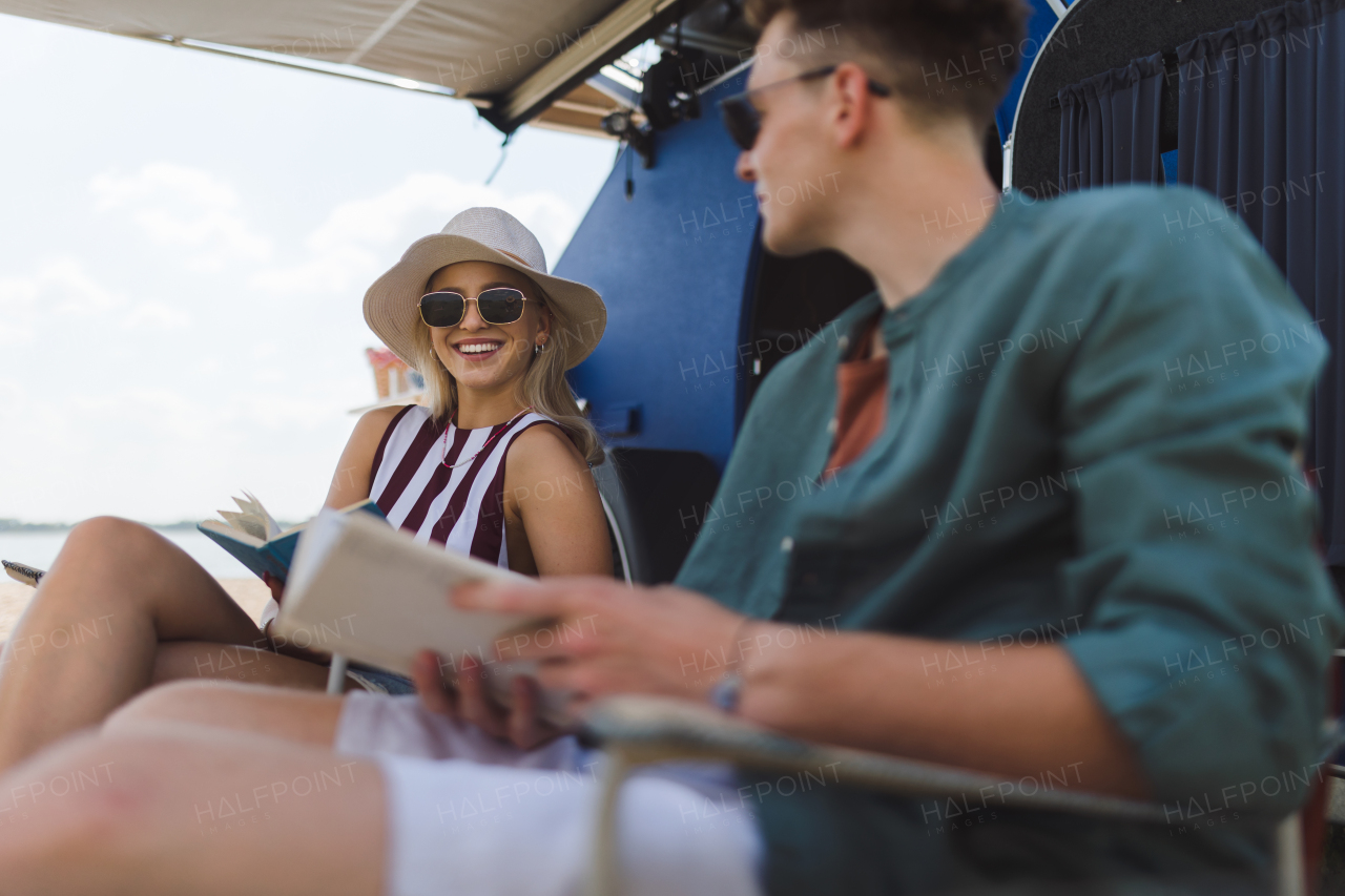 Happy young couple sitting together in front of van, camping and reading a book.