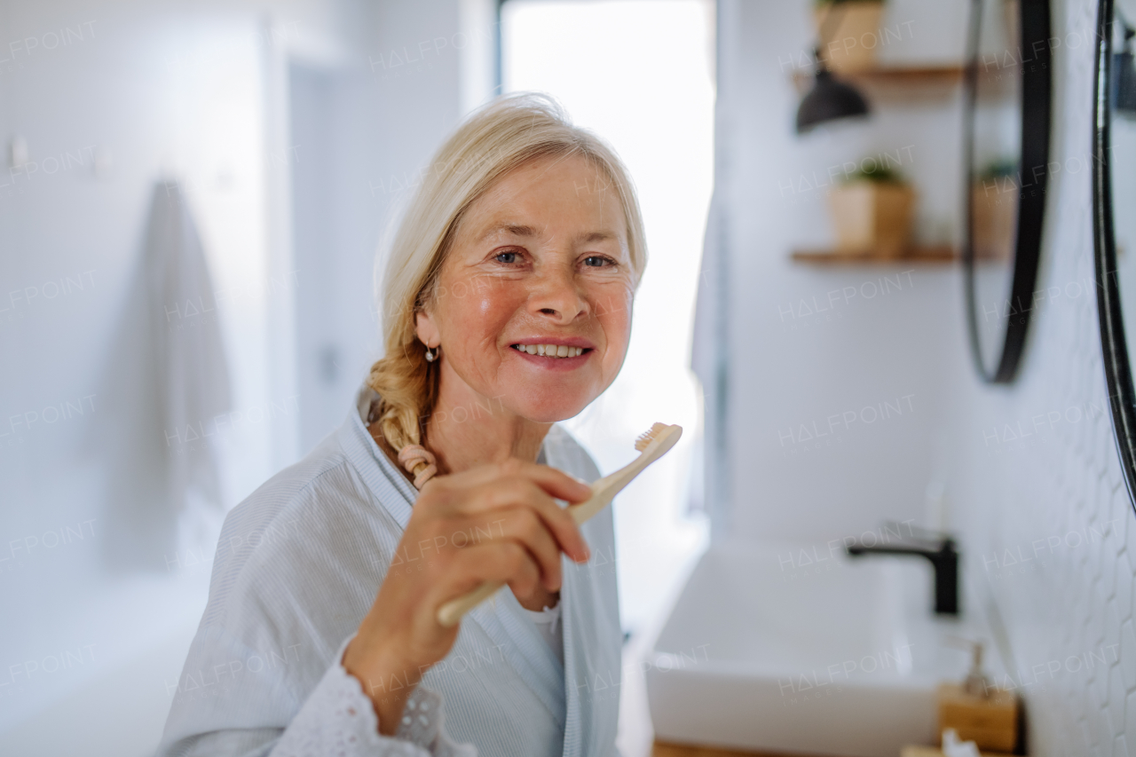 A b eautiful senior woman in bathrobe brushing teeth with eco wooden toothbrush inbathroom, sustainable lifestyle.