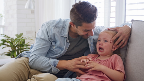 A young father sitting on sofa, holding and calming down his little daughter indoors at home.