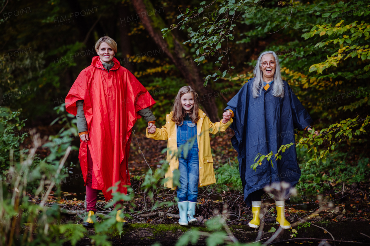 a Portrait of small girl with mother and grandmother in raincoats standing by lake outoors in nature.