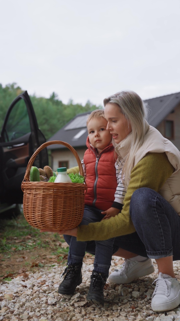 A mother and son after grocery shopping by electric car parked in front of house. House with rooftop solar panels and own EV home charging station.