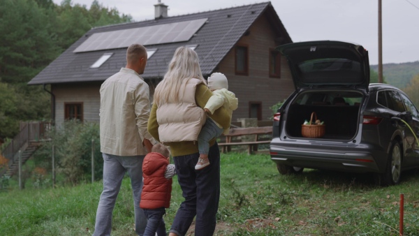 A rear view of family walking to their electric car parked in front of house. House with rooftop solar panels and own EV home charging station.