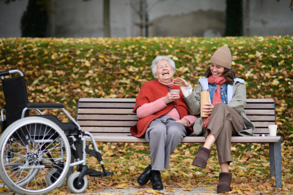 Portrait of granddaughter and grandmother sitting on bench in autumn park, drinking coffee, laughing together