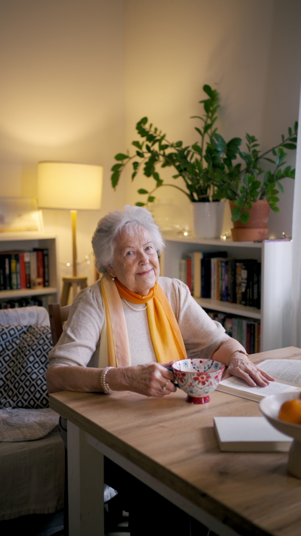Front view of elderly lady reading a book, sitting at the kitchen table and her sipping tea.