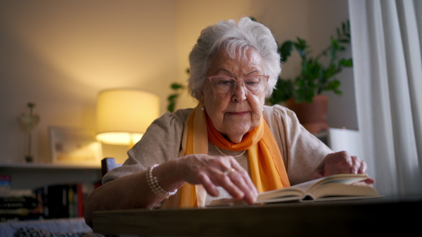 Front view of elderly lady reading a book, sitting at the kitchen table and her sipping tea.