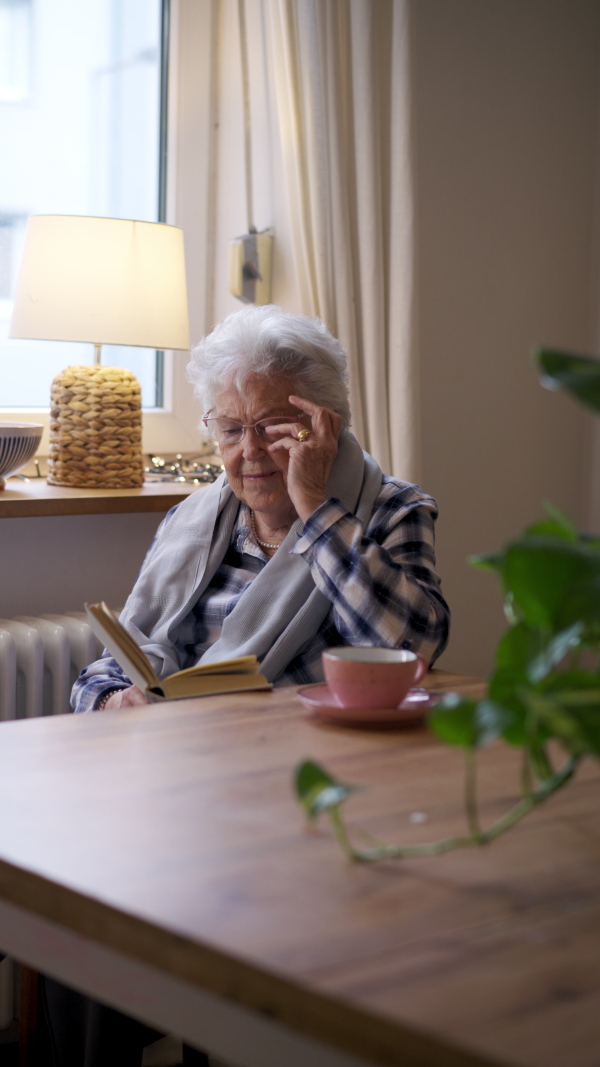 Side view of elderly lady reading a book, sitting at the kitchen table and her sipping tea.