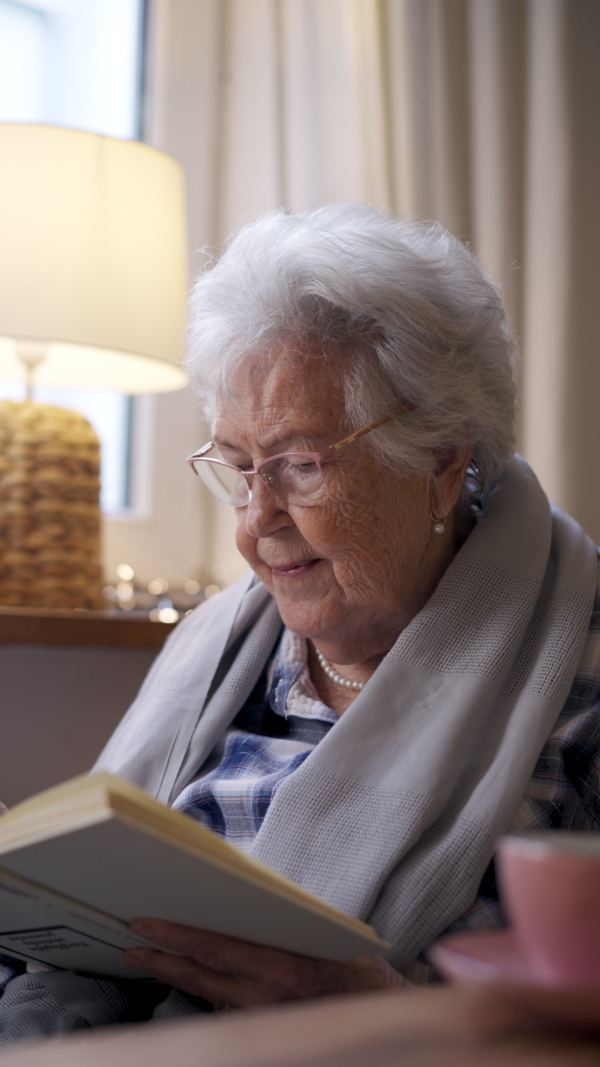 Side view of elderly lady reading a book, sitting at the kitchen table and her sipping tea.