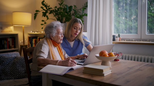 Social worker helping elderly lady with managing her finances and teaching her to use online banking on laptop.