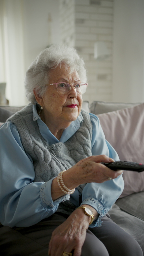 Adorable senior woman watching television, holding TV remote controller.