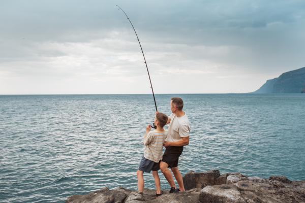 Rear view of father and young son fishing in the sea. Standing on rock holding fish rod.