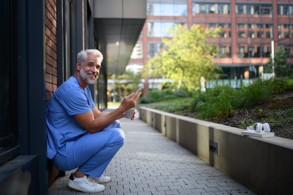 Healthcare worker in scrubs sitting outside hospital and checking phone during break.