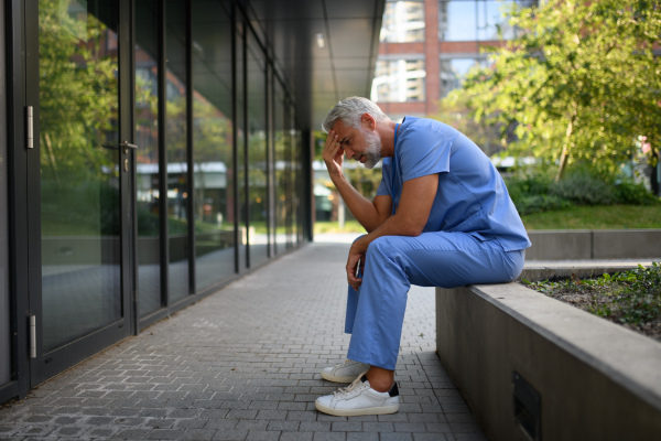 Frustrated and burnt-out doctor sitting outside the hospital, feeling down. Mental health problems.