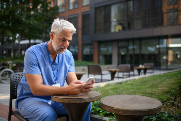 Healthcare worker in scrubs sitting outside hospital and checking phone during break.