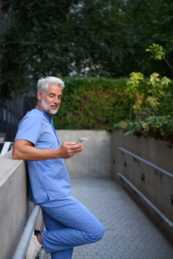 Healthcare worker in scrubs sitting outside hospital and checking phone during break.