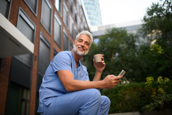 Healthcare worker in scrubs sitting outside hospital and checking phone during break.