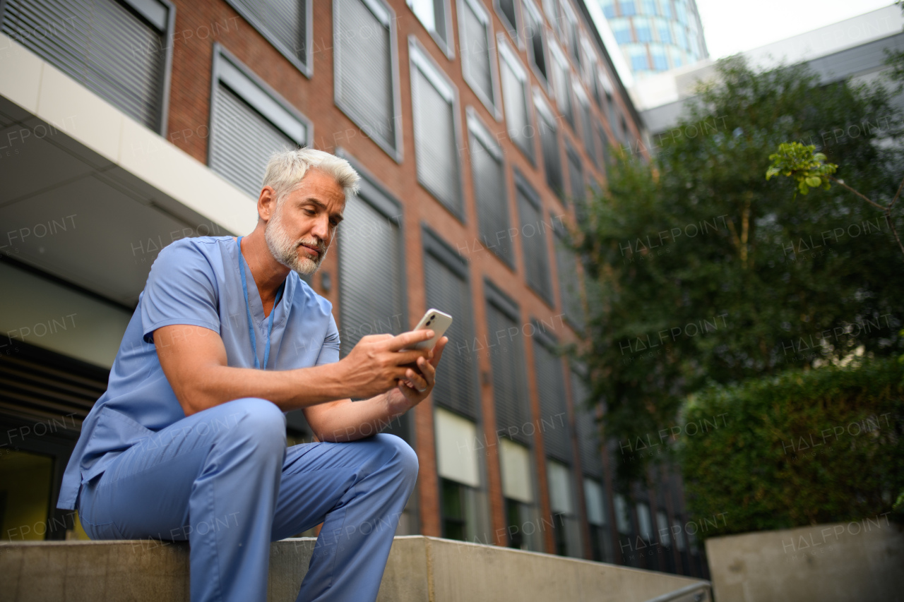 Healthcare worker in scrubs sitting outside hospital and checking phone during break.