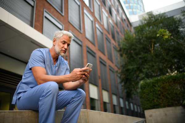 Healthcare worker in scrubs sitting outside hospital and checking phone during break.