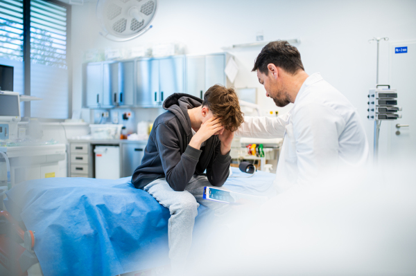 Depressed teenage boy patient sitting on hospital bed, talking with caring doctor.