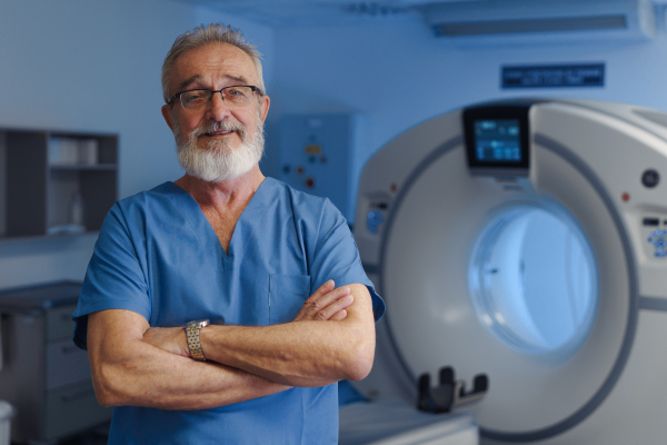 Medical specialist standing by CT scanner, looking at camera in hospital setting.
