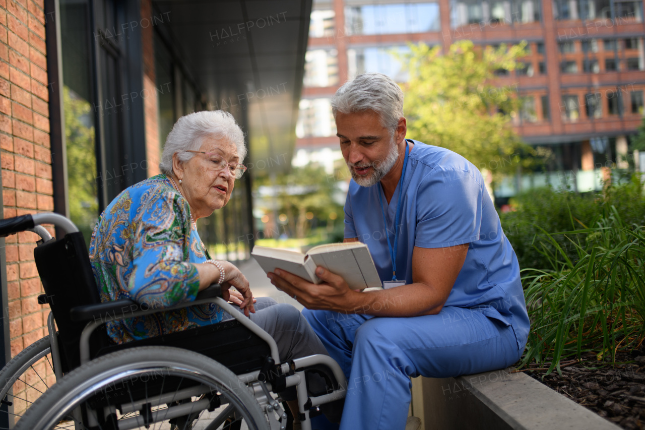 Male caregiver reading book to senior woman in a wheelchair.