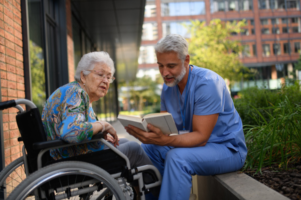 Male caregiver reading book to senior woman in a wheelchair.