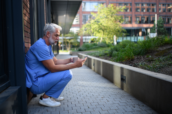 Healthcare worker in scrubs sitting outside hospital and checking phone during break.