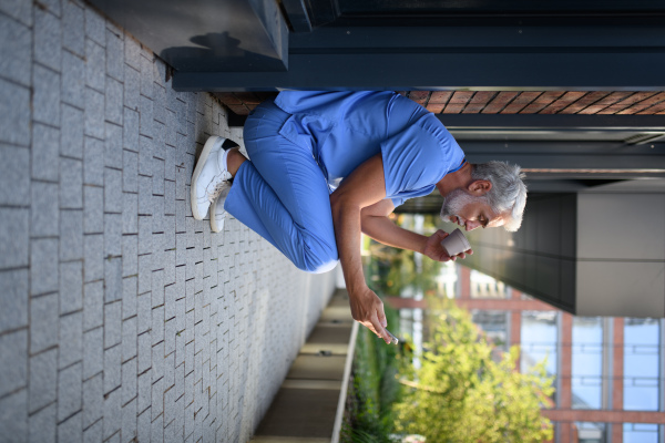 Healthcare worker in scrubs sitting outside hospital and checking phone during break.