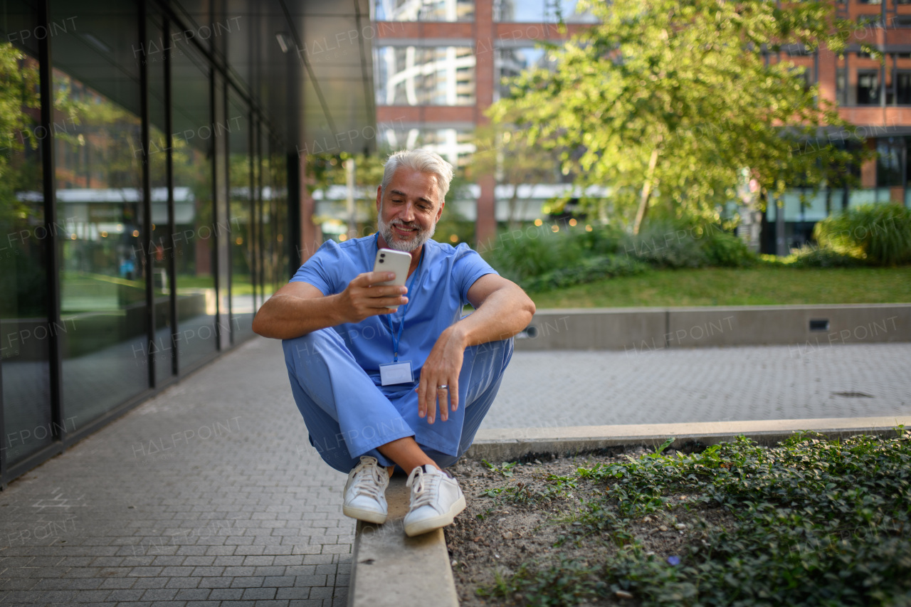 Healthcare worker in scrubs sitting outside hospital and checking phone during break.