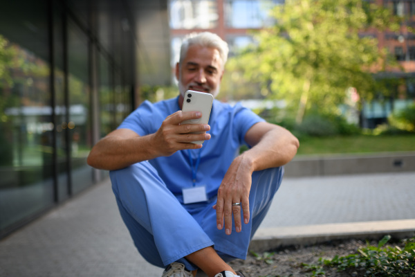 Healthcare worker in scrubs sitting outside hospital and checking phone during break.