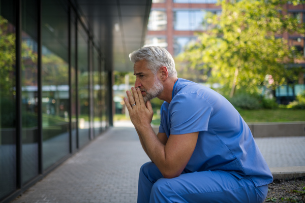 Frustrated and burnt-out doctor sitting outside the hospital, feeling down. Mental health problems.