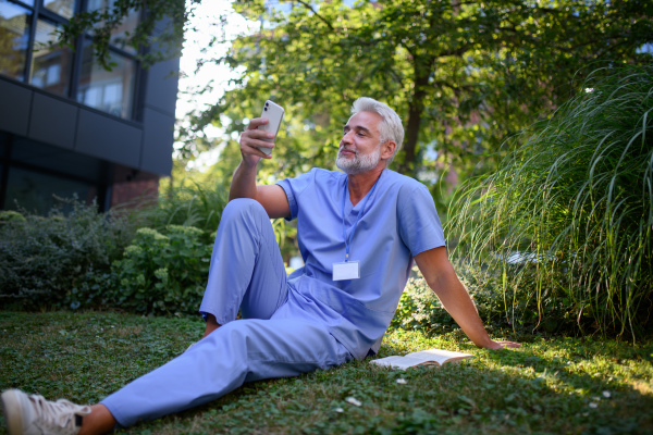 Healthcare worker resting in park outside hospital and holding smartphone.