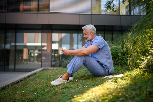 Healthcare worker resting in park outside hospital and holding smartphone.