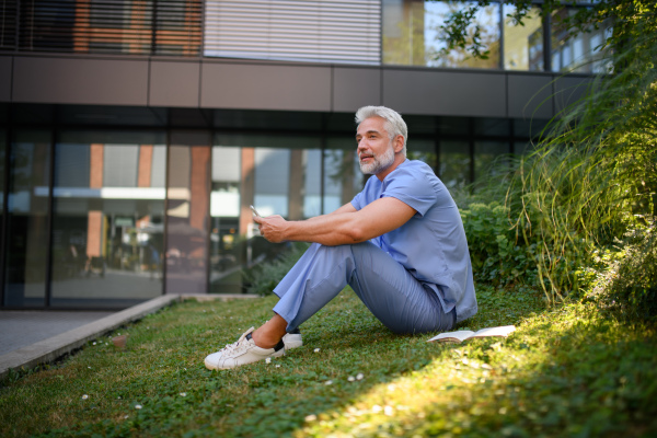 Healthcare worker resting in park outside hospital and holding smartphone.