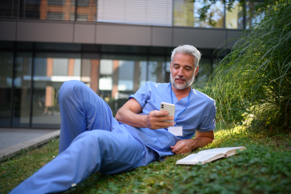 Healthcare worker resting in park outside hospital and holding smartphone.