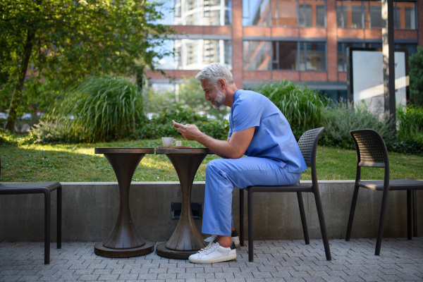 Healthcare worker in scrubs sitting outside hospital and checking phone during break.