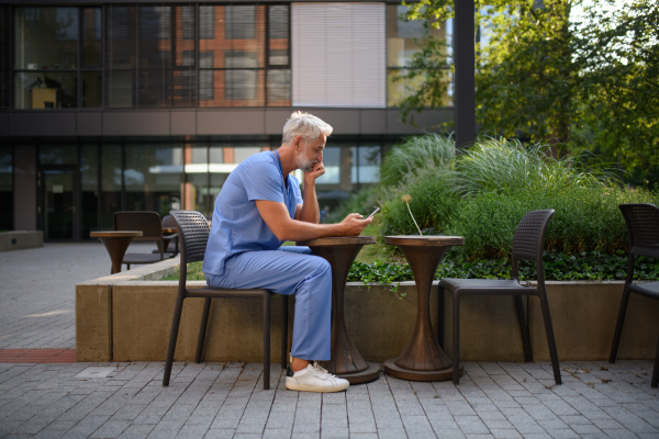 Healthcare worker in scrubs sitting outside hospital and checking phone during break.