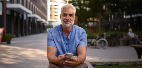 Healthcare worker in scrubs sitting outside hospital and checking phone during break. Banner with copy space.