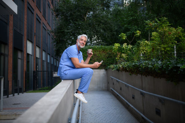 Healthcare worker in scrubs sitting outside hospital and checking phone during break.