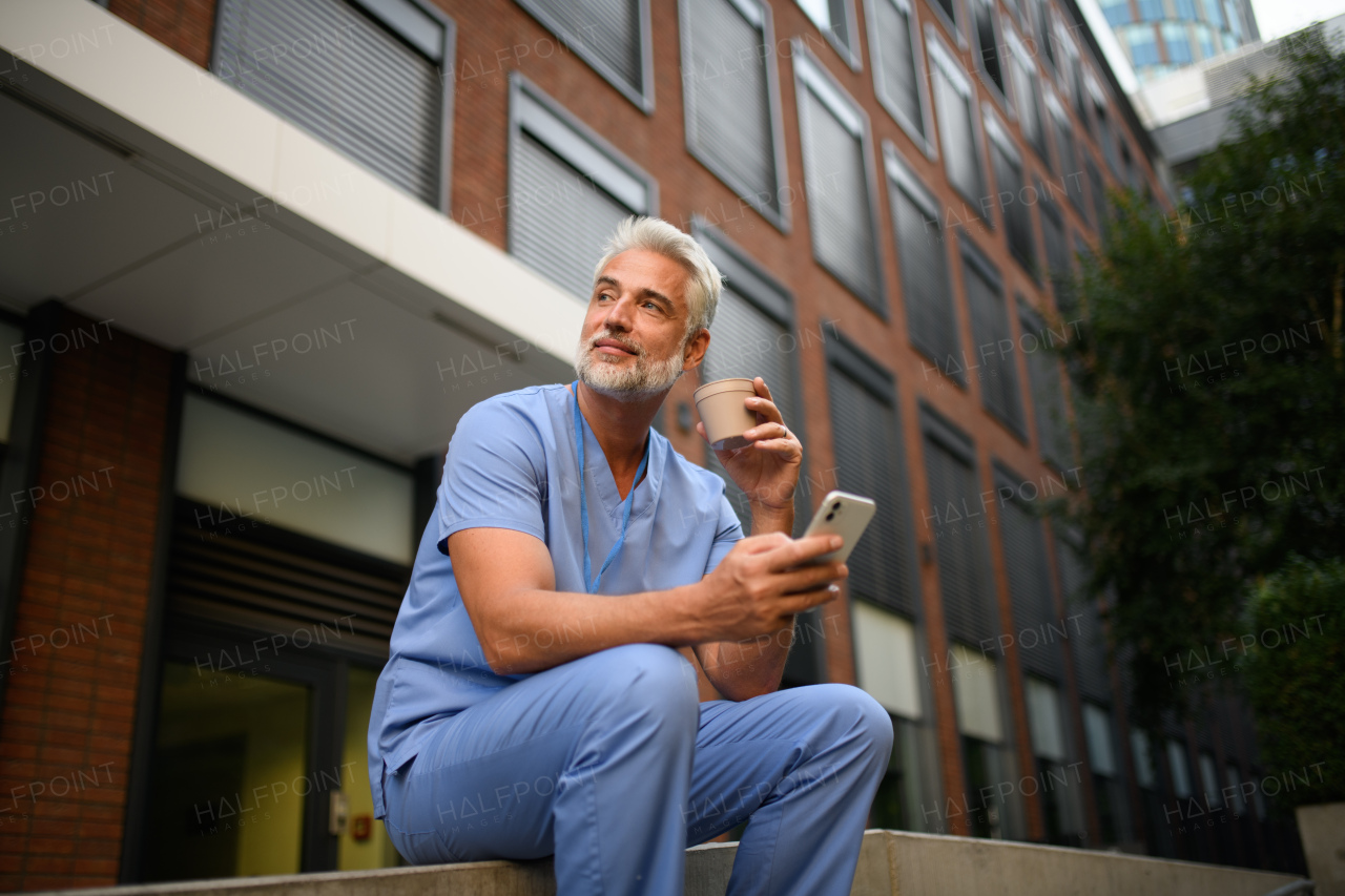Healthcare worker in scrubs sitting outside hospital and checking phone during break.