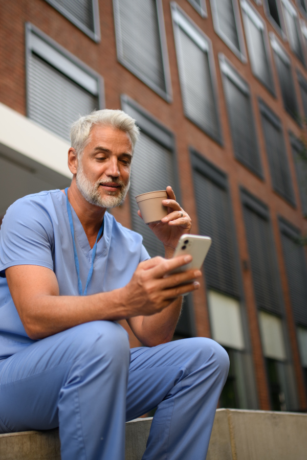 Healthcare worker in scrubs sitting outside hospital and checking phone during break.
