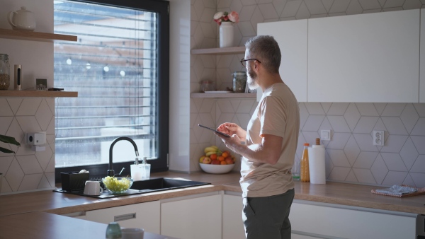 A mature man adjusting windows blind in the kitchen by tablet.