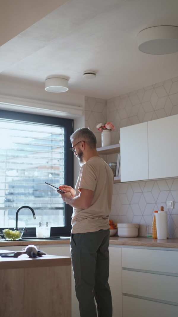 A mature man adjusting windows blind in the kitchen by tablet.