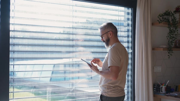A mature man adjusting windows blind in the kitchen by tablet.