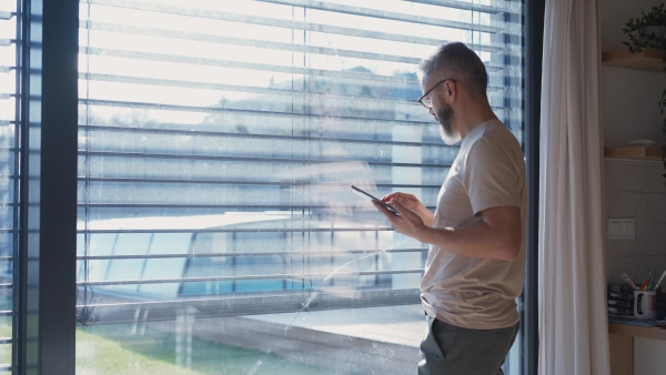 A mature man adjusting windows blind in the kitchen by tablet.