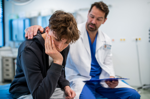 Depressed teenage boy patient sitting on hospital bed, talking with caring doctor.