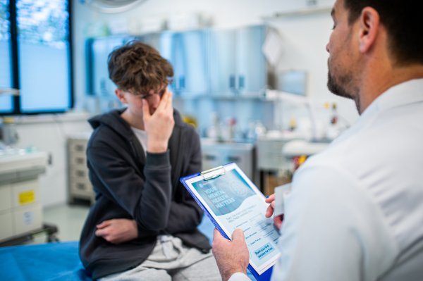 Depressed teenage boy patient sitting on hospital bed, talking with caring doctor.
